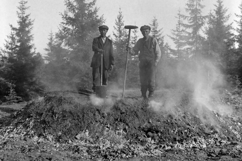 Kaksi miestä seisoo savuavan tervahaudan päällä, taustalla nuorta kuusimetsää.