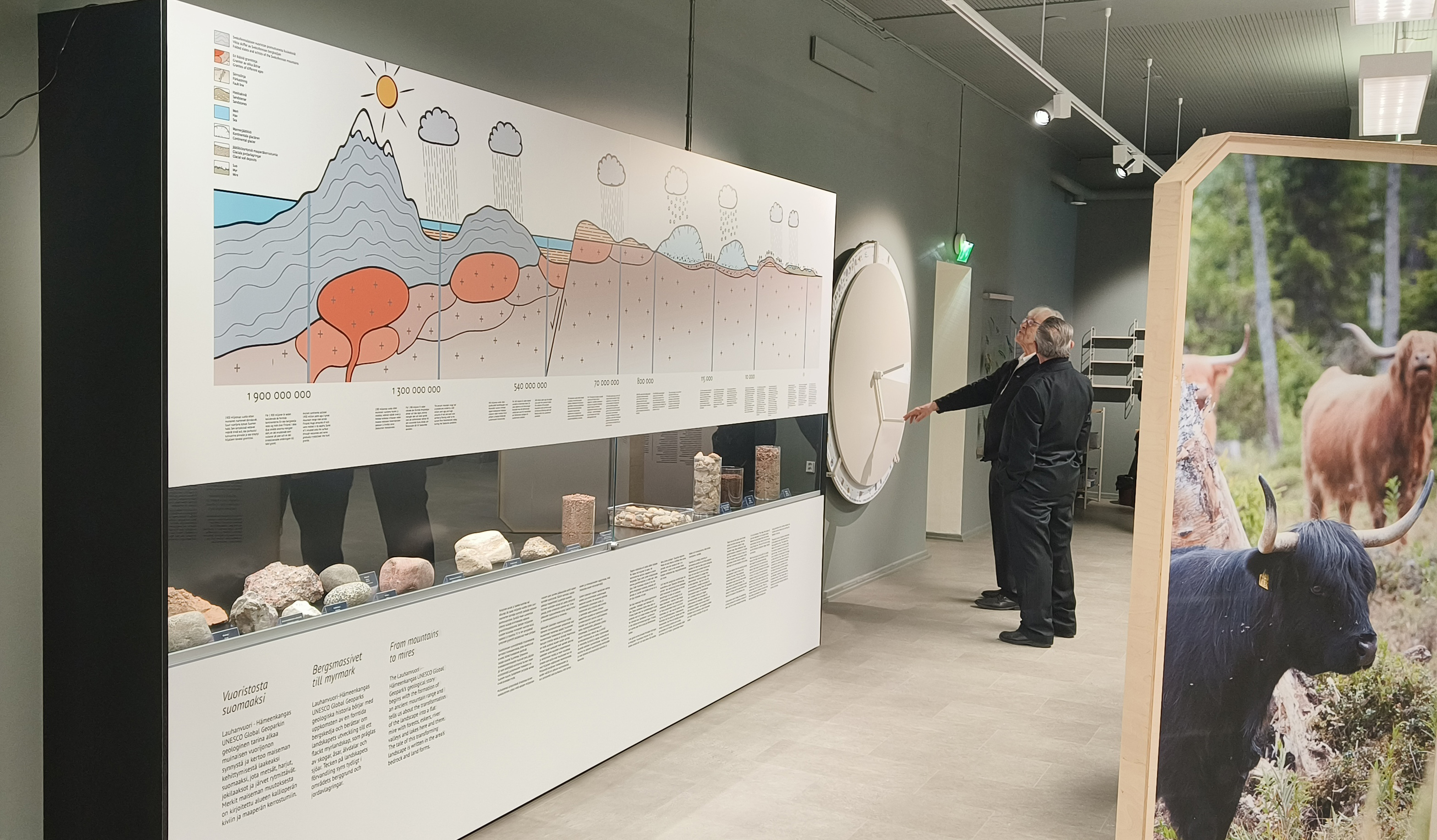 Kaksi miestä tutustumassa Geopark-näyttelyyn. Etualalla kivinäyttely upotettuna Geoparkin geologiseen aikajanaan